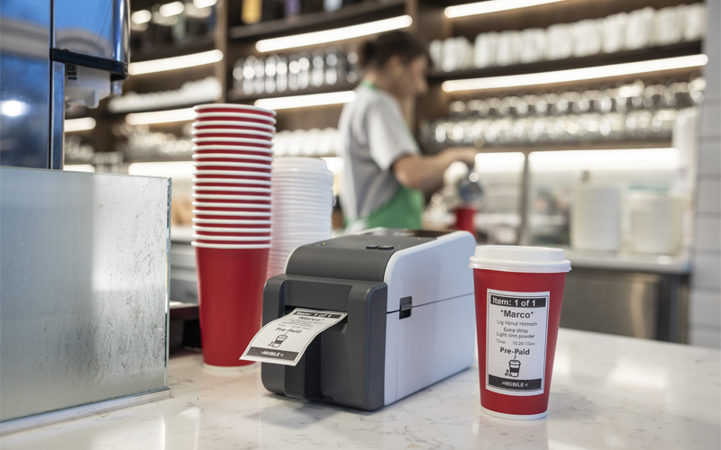 Coffee shop with woman in the background and label printer and coffee cups stacked high in the foreground